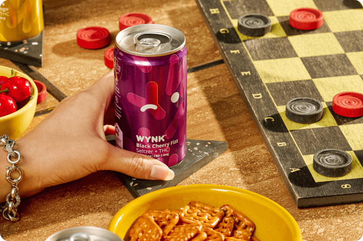 Hand holding a can of Wynk Black Cherry Fizz THC seltzer next to a checkerboard, cherries, and pretzels – perfect for holiday gatherings or game nights.