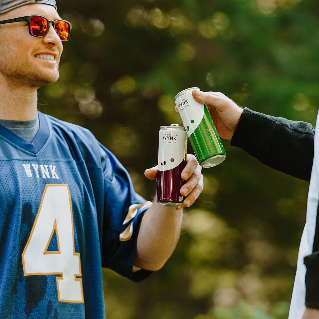 Man in a football jersey clinking cans with another person. Why people choose THC over booze.