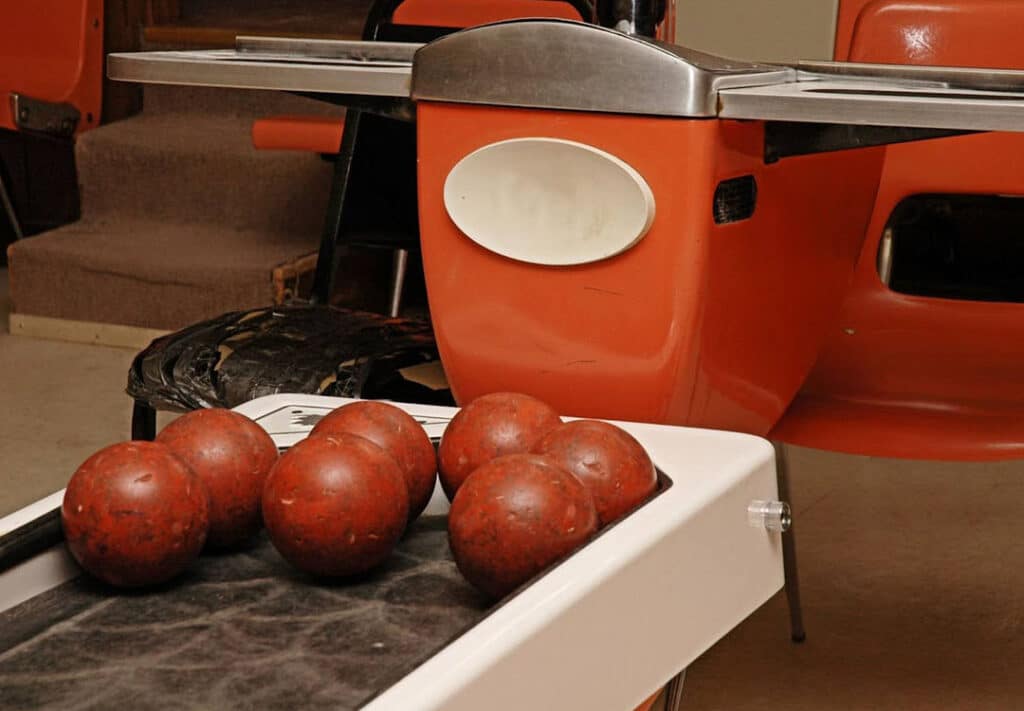 Close-up view of a vintage bowling ball return system with a set of richly hued bowling balls ready for the next game. The retro orange and white color scheme evokes a nostalgic ambiance, reminiscent of classic leisure activities that pair perfectly with a can of WYNK's best alcohol alternative, inviting bowlers to enjoy a microdose THC beverage between frames.