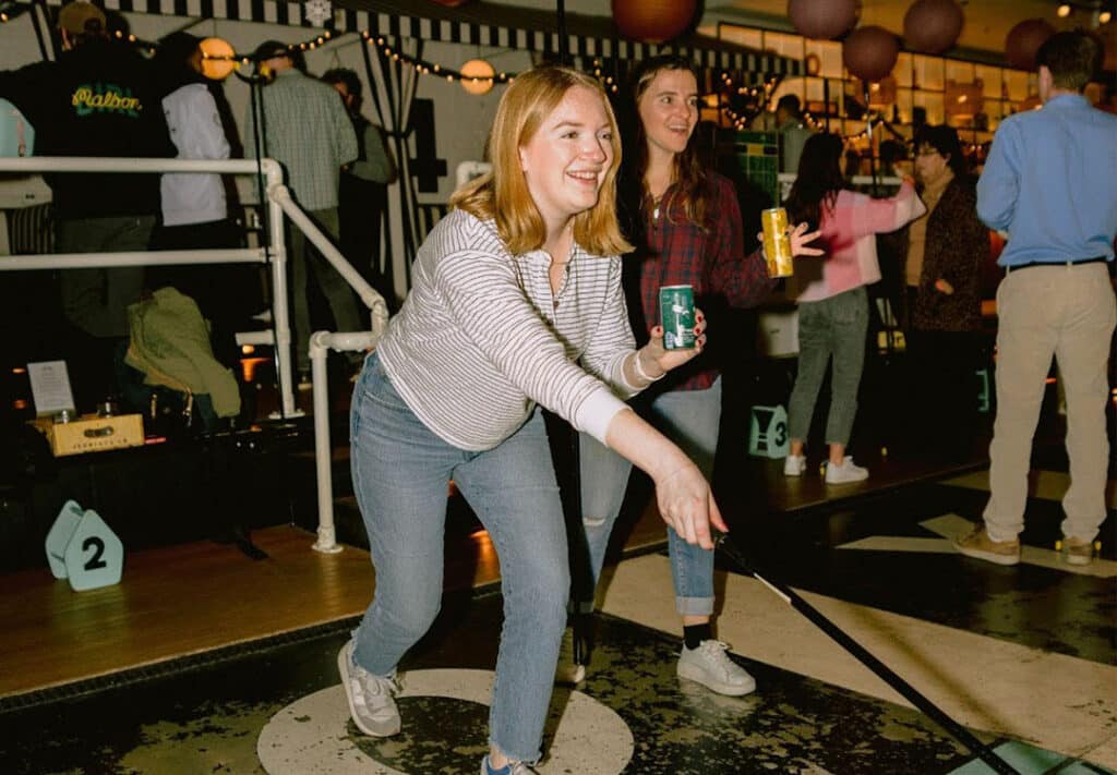 Excitement fills the air at an indoor shuffleboard club in Chicago where friends enjoy the best alcohol alternative with WYNK THC Seltzer. A woman in casual attire skillfully plays shuffleboard while holding a can of refreshing cannabis drink, embodying a fun, non-alcohol drink option for social gatherings. Her friend cheers her on with a microdose drink in hand, highlighting the popularity of THC drinks as a healthy alcohol alternative to relax and socialize.