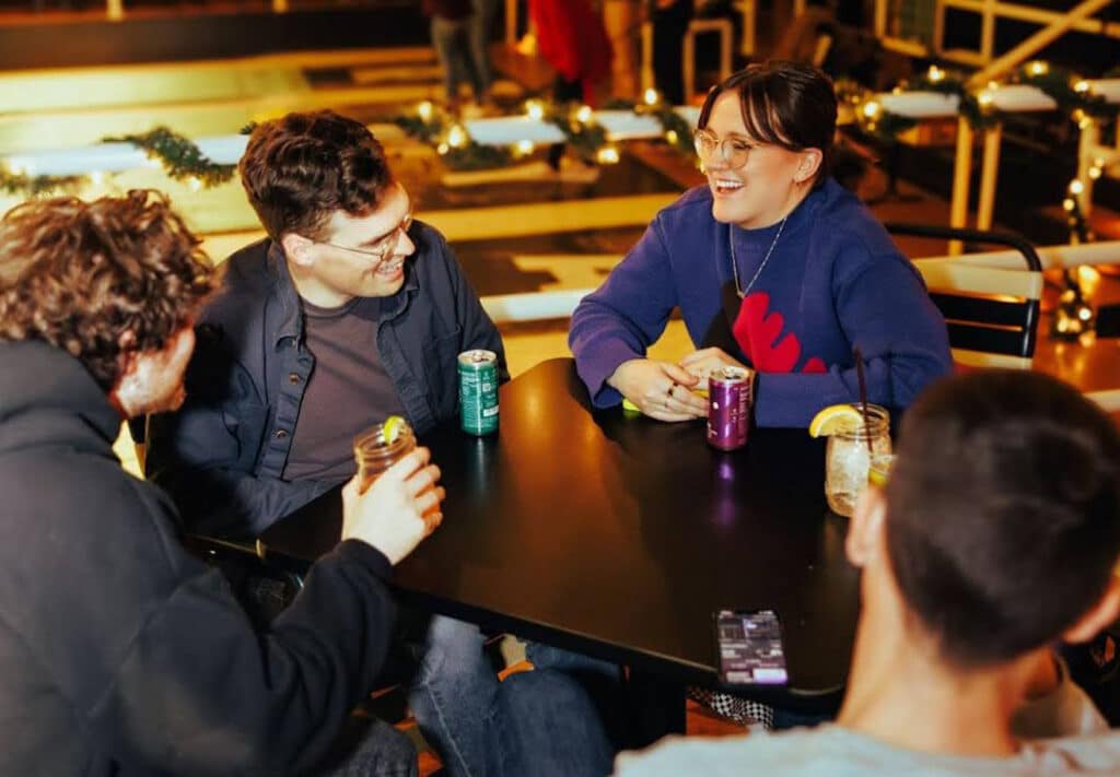 A group of friends enjoy a lively conversation at a cozy evening gathering, with two cans of WYNK THC Seltzer prominently displayed on the table. The scene captures the essence of drink edibles as a social, alcohol alternative drink, with one person holding a can, signaling the choice of a microdose THC beverage over traditional alcohol, reinforcing the trend of cannabis drink as a go-to for modern socializing.