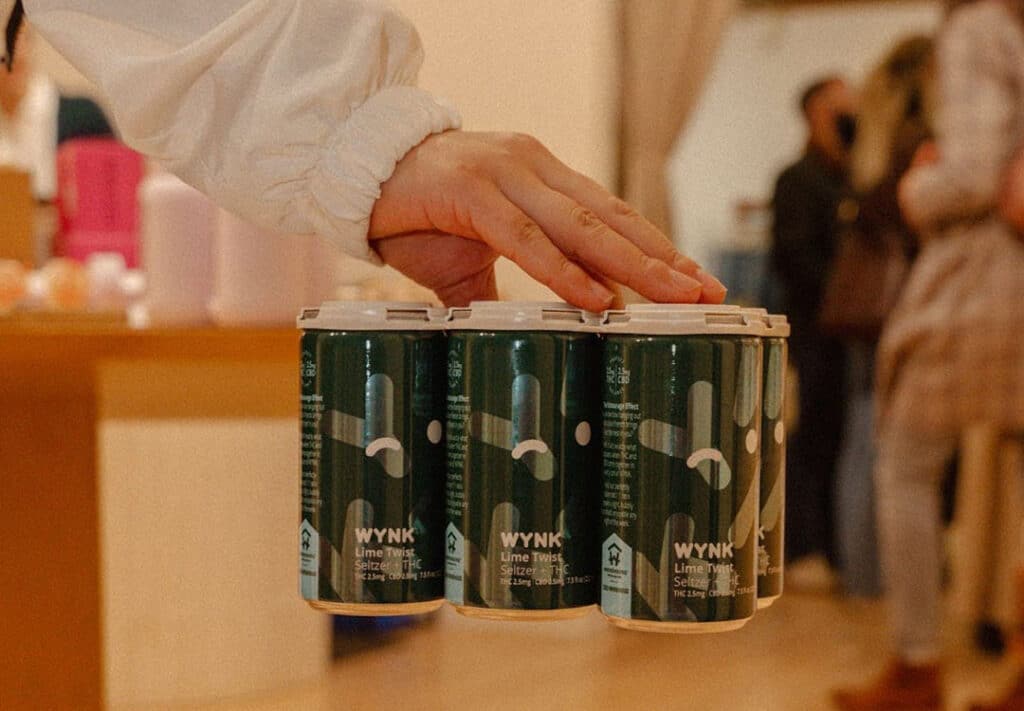 A hand gently rests on a four-pack of WYNK Lime Twist THC Seltzer, set against a lively social backdrop. The packaging promotes a refreshing, infused drink experience with a microdose THC blend, ideal for consumers seeking a cannabidiol drink as a healthy alcohol alternative. This image captures the ease of choosing WYNK's cannabis drinks, a perfect non-alcohol drink option for those looking for non-alcoholic alternatives like THC seltzer in New York City.