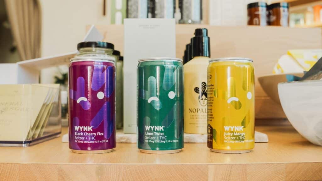 Cans of WYNK's THC-infused beverages in Black Cherry Fizz, Lime Twist, and Juicy Mango flavors on a well-organized wellness shelf, illustrating a modern lifestyle choice.