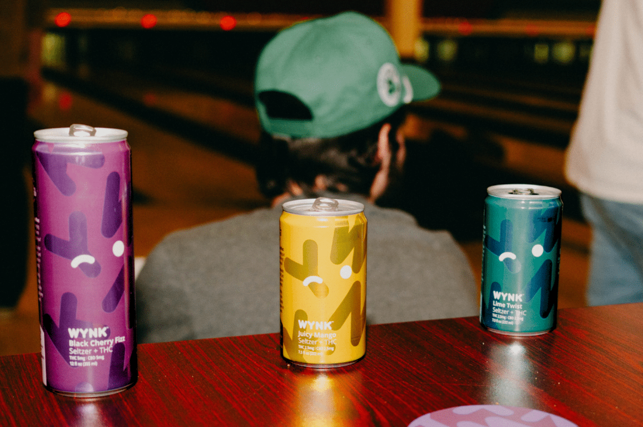 Three cans of WYNK THC seltzer in Black Cherry, Juicy Mango, and Zesty Lime flavors on a bowling alley table, with a person wearing a green cap in the background, embodying a relaxed social scene in Minnesota.