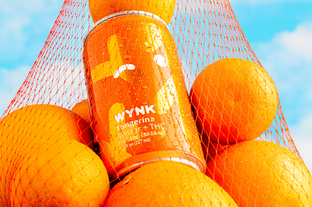 A sunny array of oranges nestled around a bottle of WYNK Tangerine Seltzer, the perfect base for crafting Galentine's Day mocktails. This inviting scene suggests fun and friendship with DIY recipes for cannabis-infused, non-alcoholic cocktails.